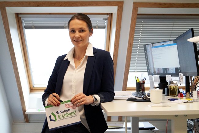 Immobilienkauffrau Carolina Opel sitzt lächelnd auf einem Schreibtischstuhl mit einem Flyer in der Hand.